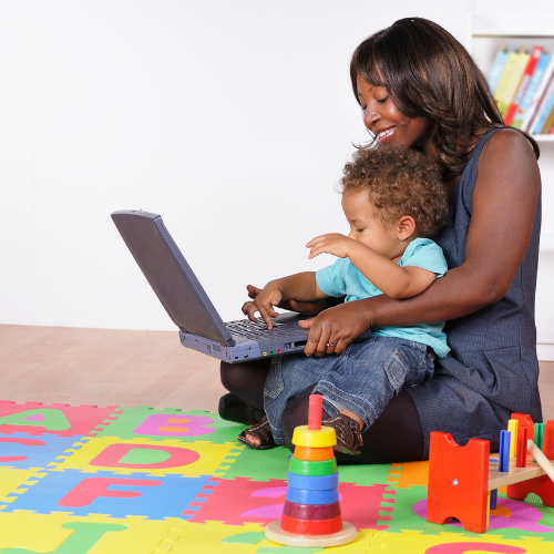 smiling woman sitting on the floor with a baby in her lap and also a laptop