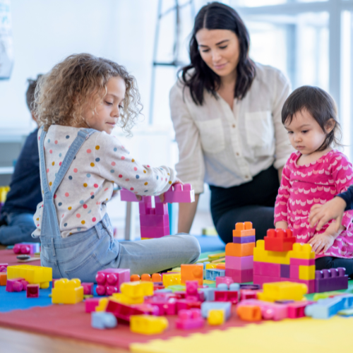 three children playing with building blocks on the floor while a female teacher overlooks